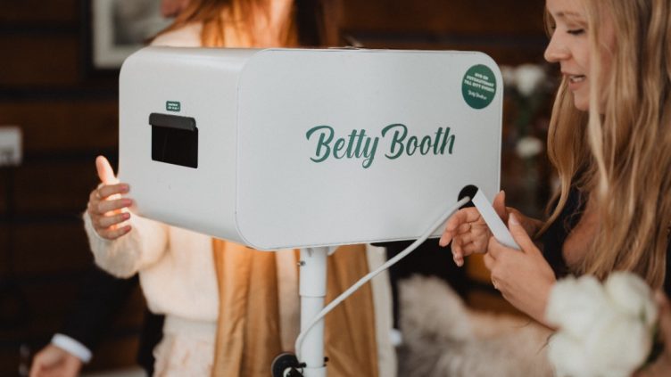 Två kvinnor interagerar med en Betty Booth fotoautomat på ett bröllop, en av dem håller i en fotoremsa som precis skrivits ut.